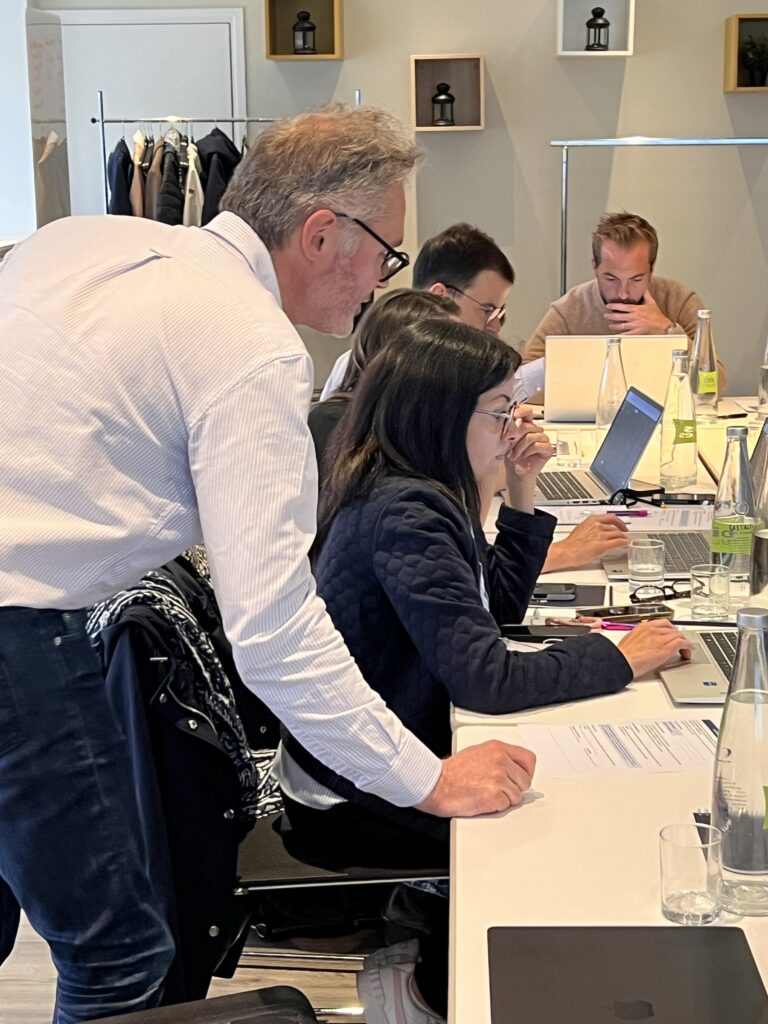 Un consultant penché vers une participante qui travaille sur son ordinateur portable, autour d’une table de travail partagée avec d’autres participants.