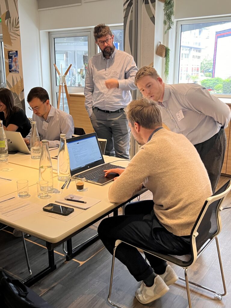 Deux consultants debout observent l’écran d’un participant assis devant son ordinateur portable, dans une salle d’atelier lumineuse.